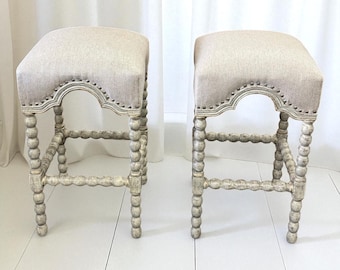May include: Two off-white, upholstered stools. Each stool has a square, light-colored fabric seat with a decorative trim. The legs are made of turned wood with a distressed, off-white finish, and a bobbin design.