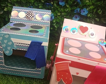 May include: Two decorative paper ovens, one blue and one pink, sit on a green surface. The blue oven has a blue and white chevron pattern, a blue ribbon, and a mitten. The pink oven has a red ribbon and a red mitten.
