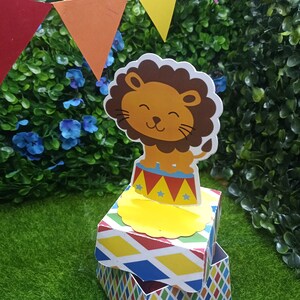 May include: A colorful, multi-layered box with a lion on top. The box is decorated with a red, yellow, blue, and green diamond pattern. The lion is brown with a yellow mane and is sitting on a red and white striped platform with yellow stars.