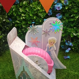 May include: A blue and white cardboard party favor box with a pink lid. The box features a cutout of a castle and a snowflake design. The box also features a character from the movie Frozen.