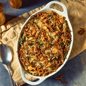 A white ceramic dish filled with a green bean casserole, topped with fried onions, sits on a beige cloth napkin. A wooden pepper grinder and a spoon are placed next to the dish, and a wooden cutting board is visible in the background.