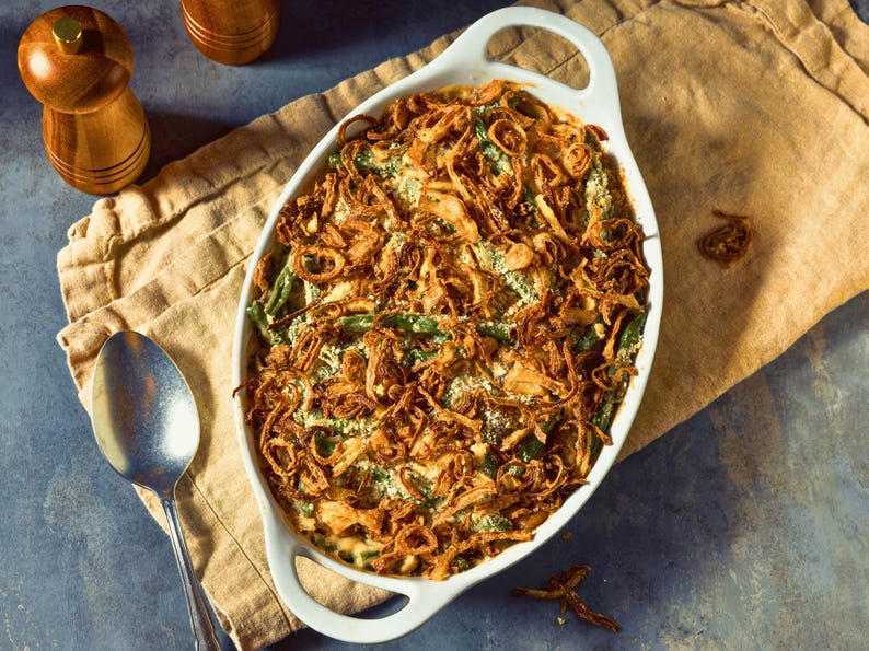 A white ceramic dish filled with a green bean casserole, topped with fried onions, sits on a beige cloth napkin. A wooden pepper grinder and a spoon are placed next to the dish, and a wooden cutting board is visible in the background.