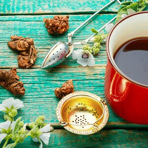 A cup of tea, a metal strainer, and various herbs and Medicinal Marshmallow flowers are arranged on a weathered wooden table.