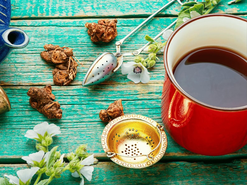 A cup of tea, a metal strainer, and various herbs and Medicinal Marshmallow flowers are arranged on a weathered wooden table.