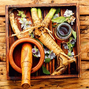 A wooden tray containing various herbs, Medicinal Marshmallow flowers, and a mortar and pestle, all arranged on a wooden surface.