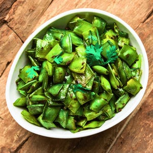 May include: A white bowl filled with sauteed green beans, garnished with fresh cilantro. The beans are a vibrant green color, with visible spices and a slightly glossy appearance. The bowl sits on a rustic wooden surface.