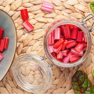 May include: A glass jar filled with chopped red and pink stalks of rhubarb. The rhubarb is sitting on a woven wicker mat. There is a plate of chopped rhubarb in the background.