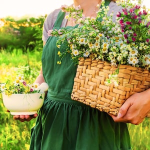 May include: A person wearing a green apron holds a wicker basket filled with a variety of wildflowers. The basket is held in the person's right hand, while their left hand holds a white mortar and pestle filled with more wildflowers.