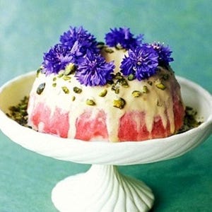 May include: A pink and white cake topped with a white icing and pistachios. The cake is decorated with purple flowers.