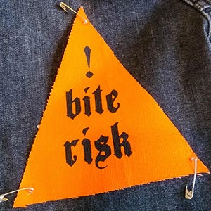 Könnte beinhalten: Dunkle Jeansjacke mit einem orangefarbenen Dreieck-Patch. Der mit Sicherheitsnadeln befestigte Patch zeigt die Worte "bite risk" in schwarzer gotischer Schrift und ein Ausrufezeichen.