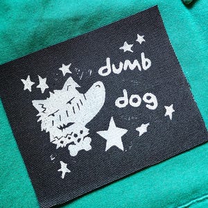 May include: A rectangular black fabric patch featuring a white screen-printed graphic of a dog head and the text "dumb dog" surrounded by small stars. The patch is sewn onto a vibrant green collared shirt with a visible chest pocket.