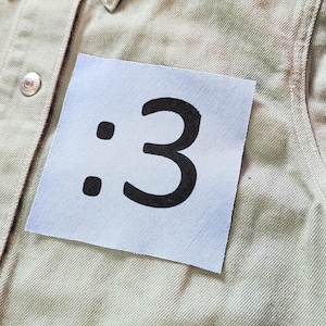 May include: A light olive green denim shirt featuring a pointed collar, silver-toned metal buttons, and a square patch on the chest printed with a black colon and the number three, forming a colon-three emoticon, with a small red Levi's tag.