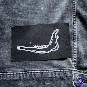 Könnte beinhalten: Dunkelgraue Jeansjacke mit einem schwarzen Aufnäher mit einer weißen Illustration eines Kieferknochens. Die Jacke hat einen Knopfverschluss und sichtbare Nahtdetails. Die Gesamtästhetik ist edgy und alternativ.