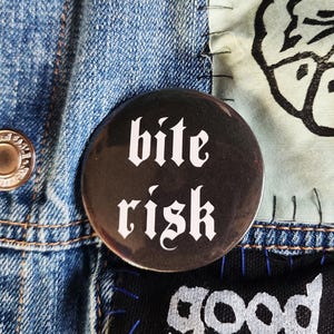 May include: A close-up of a denim jacket featuring a black button with the words "bite risk" in white gothic lettering. Patches include a skull design and the words "good bo" in white. The jacket has metal buttons.