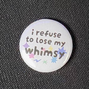 Może przedstawiać: Okrągła przypinka z białym tłem, zawierająca napis "i refuse to lose my whimsy" ciemnoszarą czcionką. Wzór jest ozdobiony małymi, kolorowymi gwiazdkami i zygzakami w odcieniach różu, żółci, błękitu oraz zieleni.