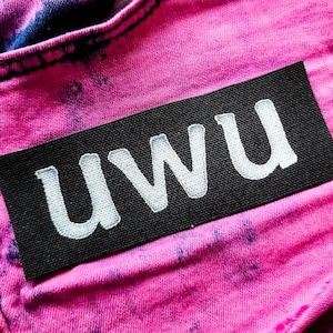 May include: A rectangular black fabric patch with the white embroidered text "uwu" stitched onto a pair of bright pink acid-wash denim jeans. The jeans feature a classic five-pocket design with a visible silver metal button at the waistline.