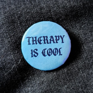 Może przedstawiać: Jasnoniebieski guzik z napisem "THERAPY IS COOL" w ciemnoniebieskich gotyckich literach. Guzik jest okrągły i umieszczony na ciemnym dżinsowym tle.