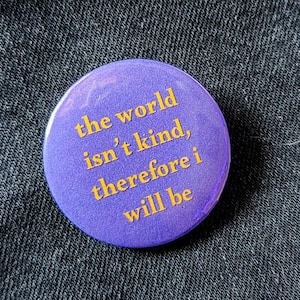 Może przedstawiać: Okrągła, fioletowa przypinka z żółtym napisem "the world isn't kind, therefore i will be". Przypinka jest umieszczona na tle ciemnego denimu.