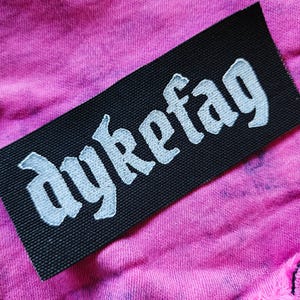 May include: A rectangular black fabric patch with the white embroidered text "dykefag" in a gothic-style font, positioned on a piece of pink acid-wash denim fabric with a frayed edge and visible stitching details.