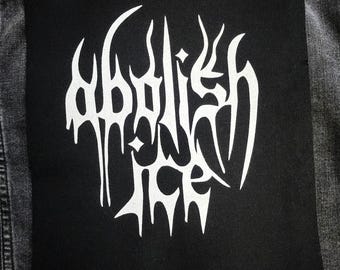 May include: A black square patch with the words "Abolish ICE" in white, stylized, gothic lettering. The patch is sewn onto a dark gray denim jacket. The image is a close-up shot.