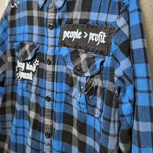 Puede incluir: Una camisa de cuadros azules y negros con un parche rectangular negro con el texto blanco people > profit, un parche más pequeño con el texto be kind be punk y varios imperdibles plateados sujetos al cuello y al bolsillo del pecho.