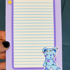 May include: A rectangular notepad with a light purple border and a cream-colored center featuring horizontal blue lines. The top left corner has yellow stars, and the bottom right corner features a blue teddy bear with a star crown.