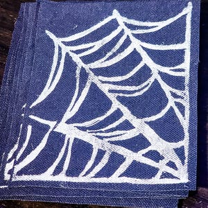 May include: A stack of square, navy blue fabric patches. Each patch features a white spiderweb design. The patches are stacked on a dark wooden surface. The image is well-lit, highlighting the contrast between the dark blue and white.