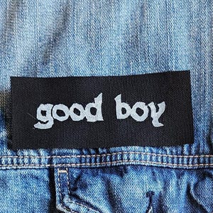 Puede incluir: Primer plano de una chaqueta vaquera azul claro con un parche negro. El parche tiene la inscripción "good boy" en blanco. La chaqueta tiene un bolsillo y botones plateados.