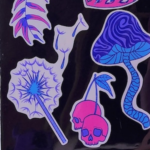 A hand with visible tattoos and blue nail polish holds the sticker pack of colorful plants against a blurred background of green trees and a wooden fence.