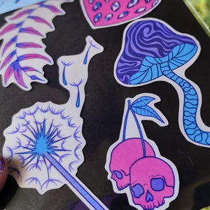 May include: A sheet of waterproof stickers with a faerie flora theme. The stickers feature illustrations of a strawberry with eyes, a mushroom, cherries with skull faces, a dandelion, and other botanical designs in shades of pink, blue, and purple.
