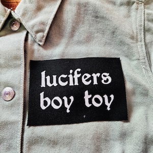 May include: A light olive green denim jacket featuring a rectangular black fabric patch sewn onto the chest. The patch displays the white embroidered text "lucifers boy toy" in a stylized font. The jacket includes silver metal buttons.