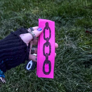 A hand with purple nail polish holds two fabric strips on grass. The black strip has a white chain design; the pink strip features a black chain. The vibe is edgy. Based on the opaque ink, you can tell the printing method is linocut.