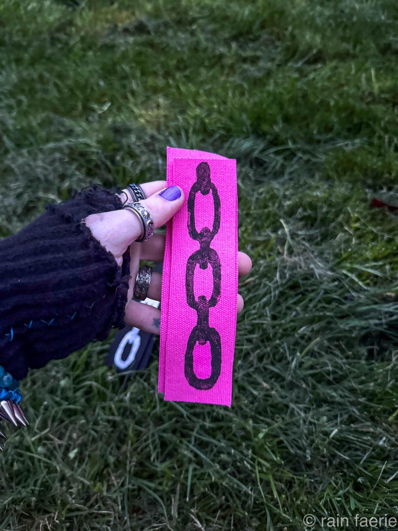 A hand with purple nail polish holds two fabric strips on grass. The black strip has a white chain design; the pink strip features a black chain. The vibe is edgy. Based on the opaque ink, you can tell the printing method is linocut.