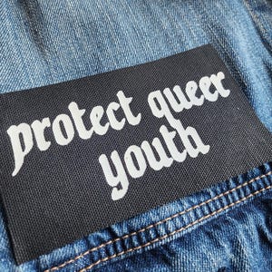 May include: A black patch with white text that reads "protect queer youth" sewn onto a blue denim jacket.