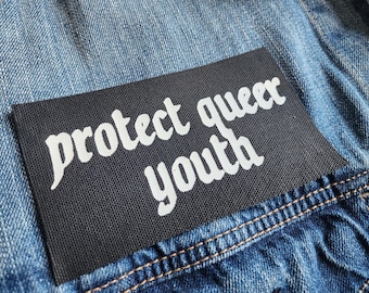 Może przedstawiać: Czarna łatka z białym tekstem, który brzmi "protect queer youth", przyszyta do niebieskiej dżinsowej kurtki.
