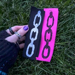 A hand with purple nail polish holds two fabric strips on grass. The black strip has a white chain design; the pink strip features a black chain. The vibe is edgy. Based on the opaque ink, you can tell the printing method is linocut.