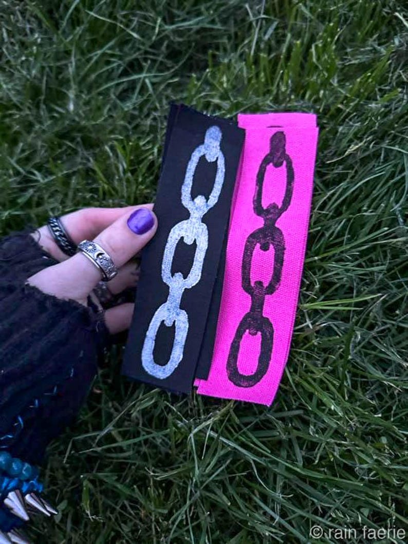A hand with purple nail polish holds two fabric strips on grass. The black strip has a white chain design; the pink strip features a black chain. The vibe is edgy. Based on the opaque ink, you can tell the printing method is linocut.