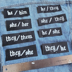 May include: A blue denim jacket adorned with black patches, each bearing white text. The patches display pronoun combinations such as "he/him", "she/her", and "they/them", alongside others. A metal ruler is positioned at the bottom of the image.