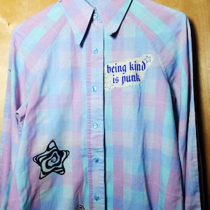 Puede incluir: Una camisa de manga larga con botones y un patrón de cuadros en azul claro y rosa. La camisa incluye un parche de tela blanco con el texto azul "being kind is punk" y dibujos en tinta negra de una estrella en espiral y un pequeño hongo.
