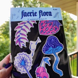 A hand with visible tattoos and blue nail polish holds the sticker pack of colorful plants against a blurred background of green trees and a wooden fence.