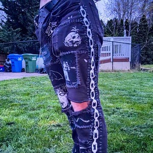 A person wears black punk-style pants with graphic patches, standing to the side showing off the chain patch running down the entire pant leg. They have one knee cutout, and are standing on grass. The style is edgy and rebellious.