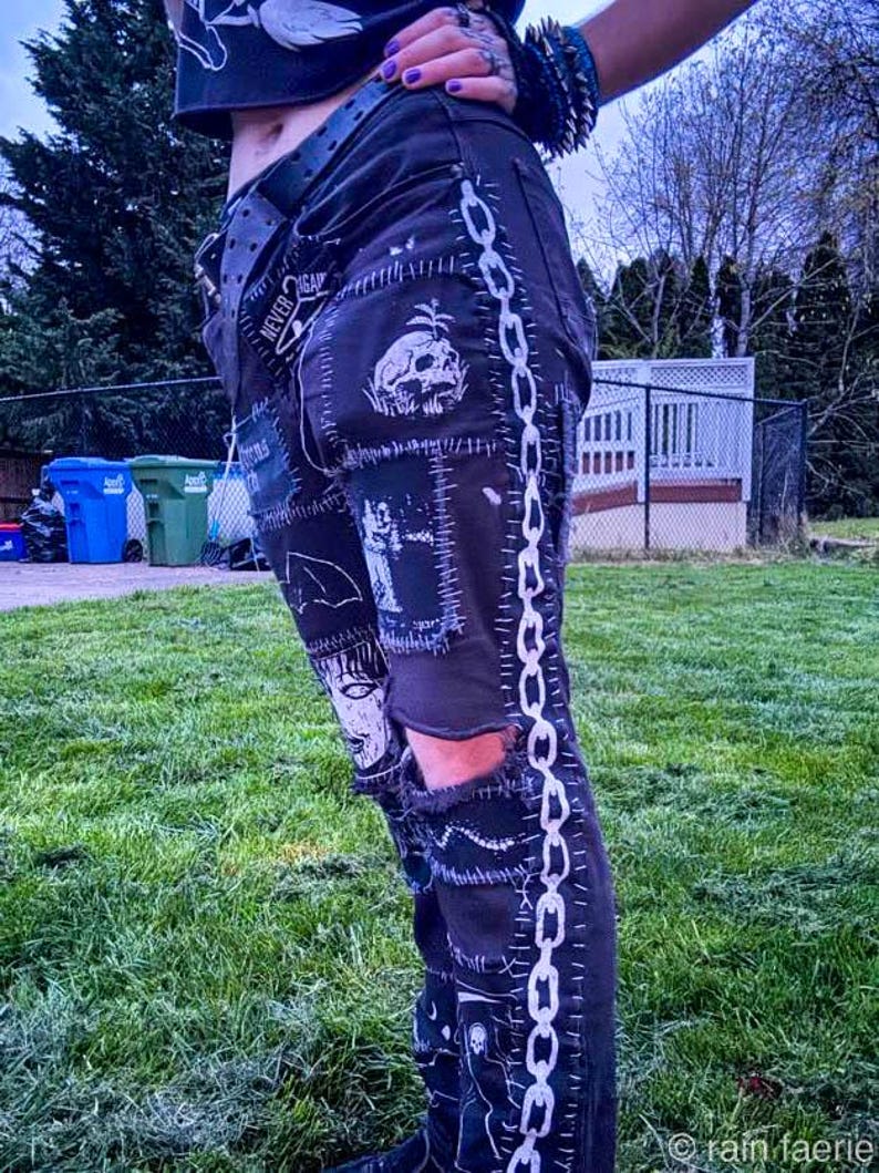 A person wears black punk-style pants with graphic patches, standing to the side showing off the chain patch running down the entire pant leg. They have one knee cutout, and are standing on grass. The style is edgy and rebellious.
