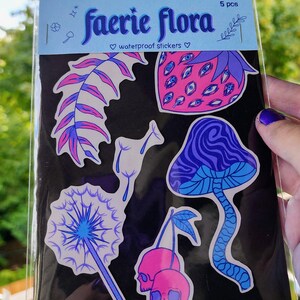 A hand with visible tattoos and blue nail polish holds the sticker pack of colorful plants against a blurred background of green trees and a wooden fence.