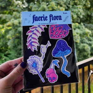 A hand with visible tattoos and blue nail polish holds the sticker pack of colorful plants against a blurred background of green trees and a wooden fence.