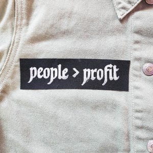 May include: A rectangular black fabric patch with white embroidered text that reads people > profit, attached to a light beige denim jacket featuring silver metal buttons and a visible collar and seam detailing on the front of the garment.