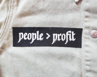 May include: A rectangular black fabric patch with white embroidered text that reads people > profit, attached to a light beige denim jacket featuring silver metal buttons and a visible collar and seam detailing on the front of the garment.