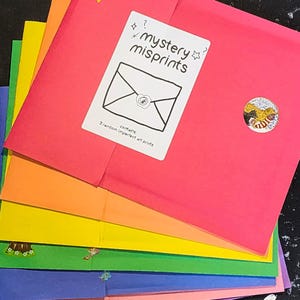 May include: A stack of colorful envelopes in a rainbow of hues, including pink, orange, yellow, green, blue, and purple. The top envelope is pink and features a sticker that says "mystery misprints" with an envelope graphic. Two yellow star stickers are also visible.