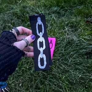 A hand with purple nail polish holds two fabric strips on grass. The black strip has a white chain design; the pink strip features a black chain. The vibe is edgy. Based on the opaque ink, you can tell the printing method is linocut.