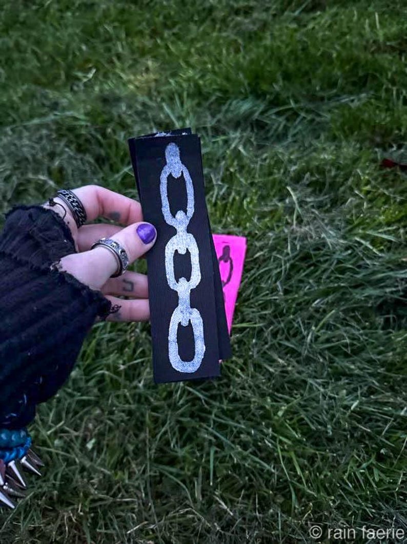 A hand with purple nail polish holds two fabric strips on grass. The black strip has a white chain design; the pink strip features a black chain. The vibe is edgy. Based on the opaque ink, you can tell the printing method is linocut.