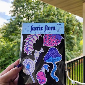 A hand with visible tattoos and blue nail polish holds the sticker pack of colorful plants against a blurred background of green trees and a wooden fence.